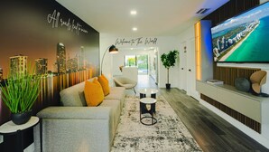 Ein modernes Wohnzimmer mit Stadtbild-Wandgemälde, einer Couch mit orangefarbenen Kissen, einem Couchtisch und einem Fernseher, der eine Strandansicht zeigt.