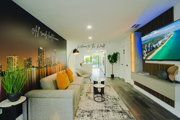 A modern living room with a cityscape mural, a sofa with orange cushions, a coffee table, and a TV displaying a beach scene.