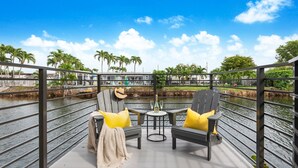 Eine Terrasse mit zwei Stühlen und einem Tisch, mit Blick auf ein Gewässer, das von Palmen und Gebäuden im Hintergrund gesäumt wird.