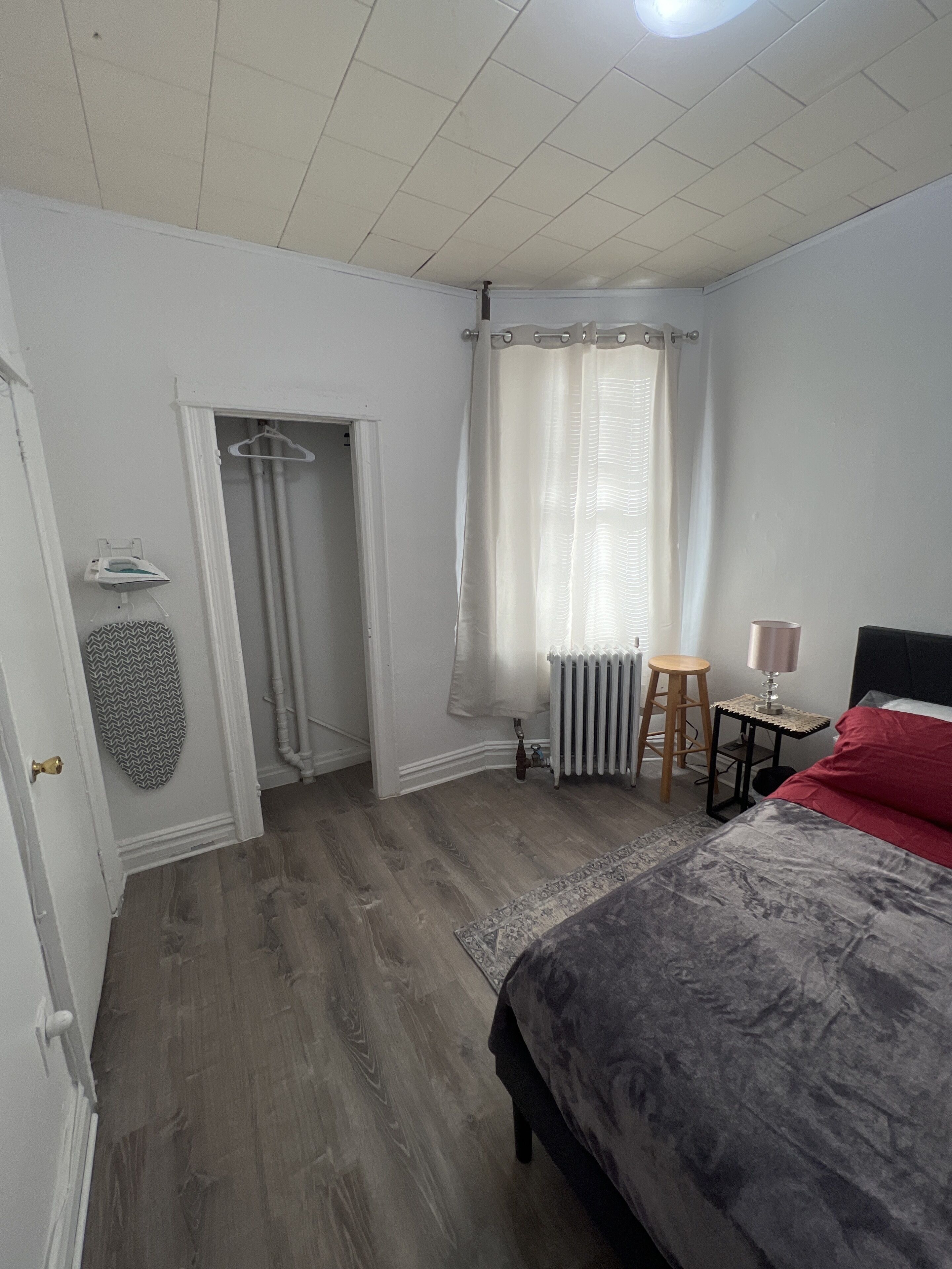 A bedroom with a bed, a radiator, a small table with a lamp, a hanging garment, and a door leading to another room.