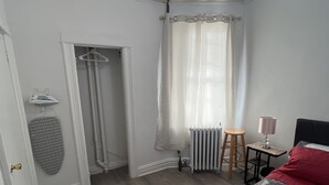 A bedroom with a bed, a radiator, a small table with a lamp, a hanging garment, and a door leading to another room.