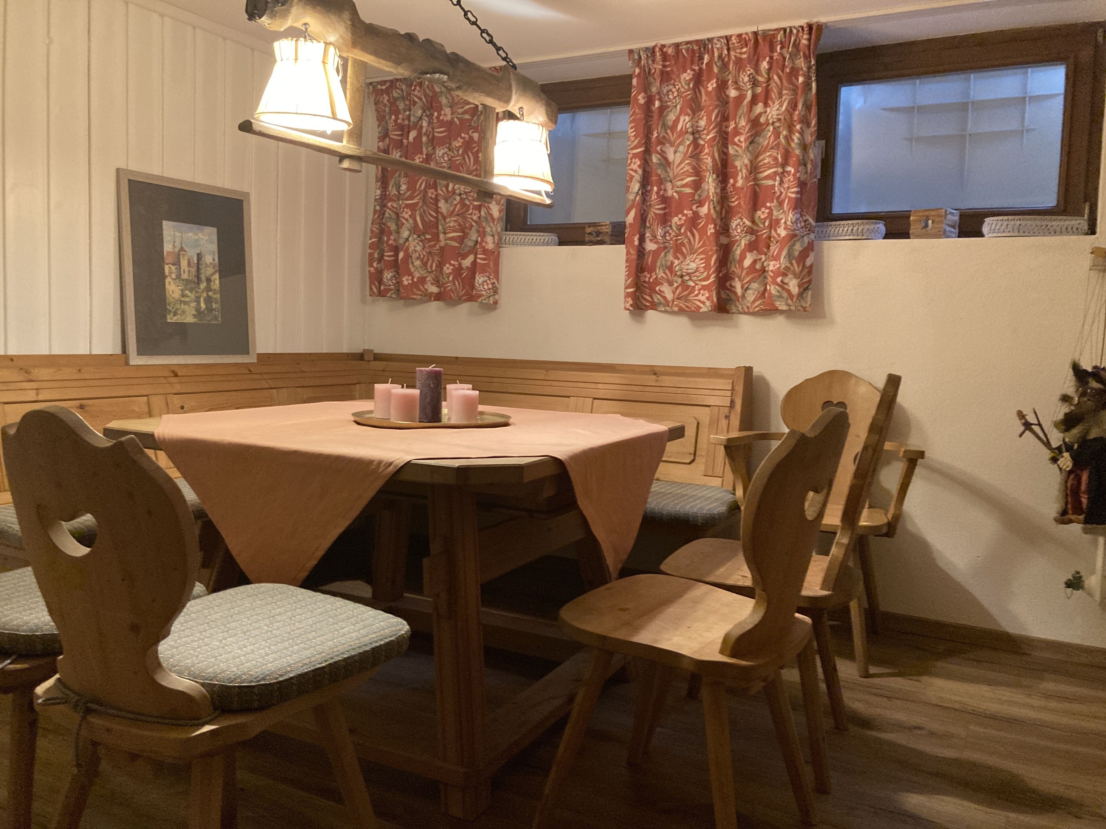 A dining area with a round table, wooden chairs, and a bench. There are candles on the table, and the room has floral curtains and a framed picture on the wall.