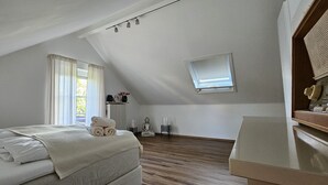 Un dormitorio con techo inclinado, una cama con ropa de cama blanca, una mesita de noche pequeña con una lámpara y una ventana con cortinas.