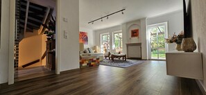 Ein geräumiger Wohnbereich mit Holzboden, einem bunten Spielhäuschen, einer Couch und einem großen Fenster mit Blick ins Freie.