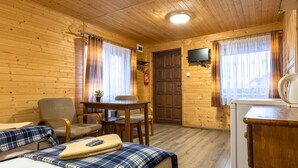 A cozy room with wooden walls, a bed with a plaid blanket, a table with chairs, a television, and a kitchenette.