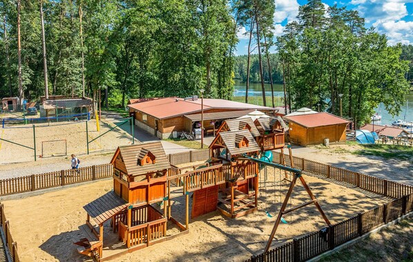 A playground with wooden play structures, a sandy area, and a building with a red roof.