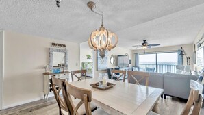 A dining area with a wooden table, chairs, and a chandelier. There is a sofa and a window with a view. - Sand Dollar #308 Beachfront 3 bedroom (Indian Shores)