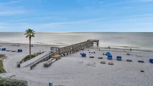 A beach with a wooden pier, blue beach umbrellas, and a palm tree. - Sand Dollar #308 Beachfront 3 bedroom (Indian Shores)