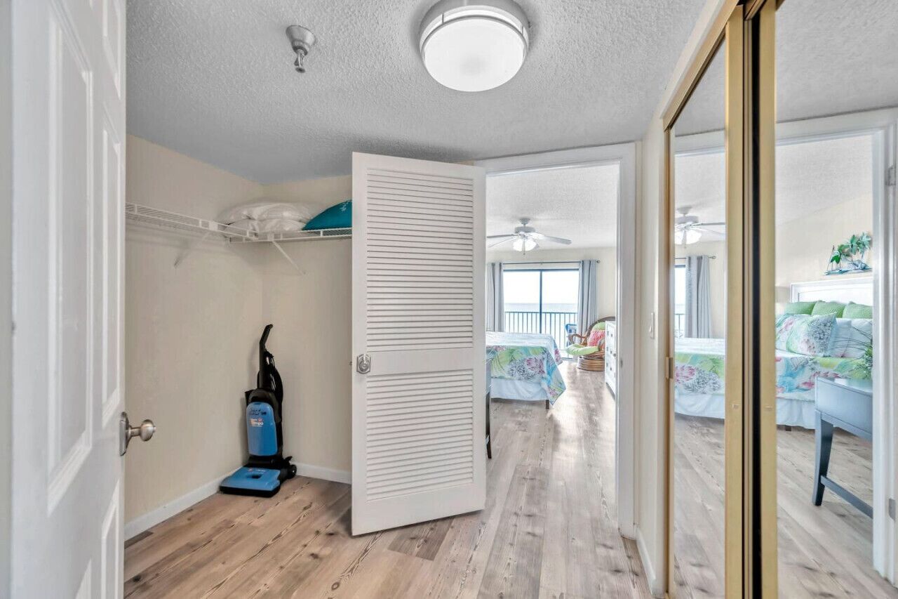 A room with a wooden floor, a ceiling fan, a bed with a floral bedspread, and a view of a balcony with a chair and plants.