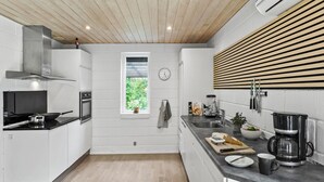 Una cocina moderna con armarios blancos, horno empotrado, estufa, ventana con vistas a zonas verdes, un reloj y una cafetera.