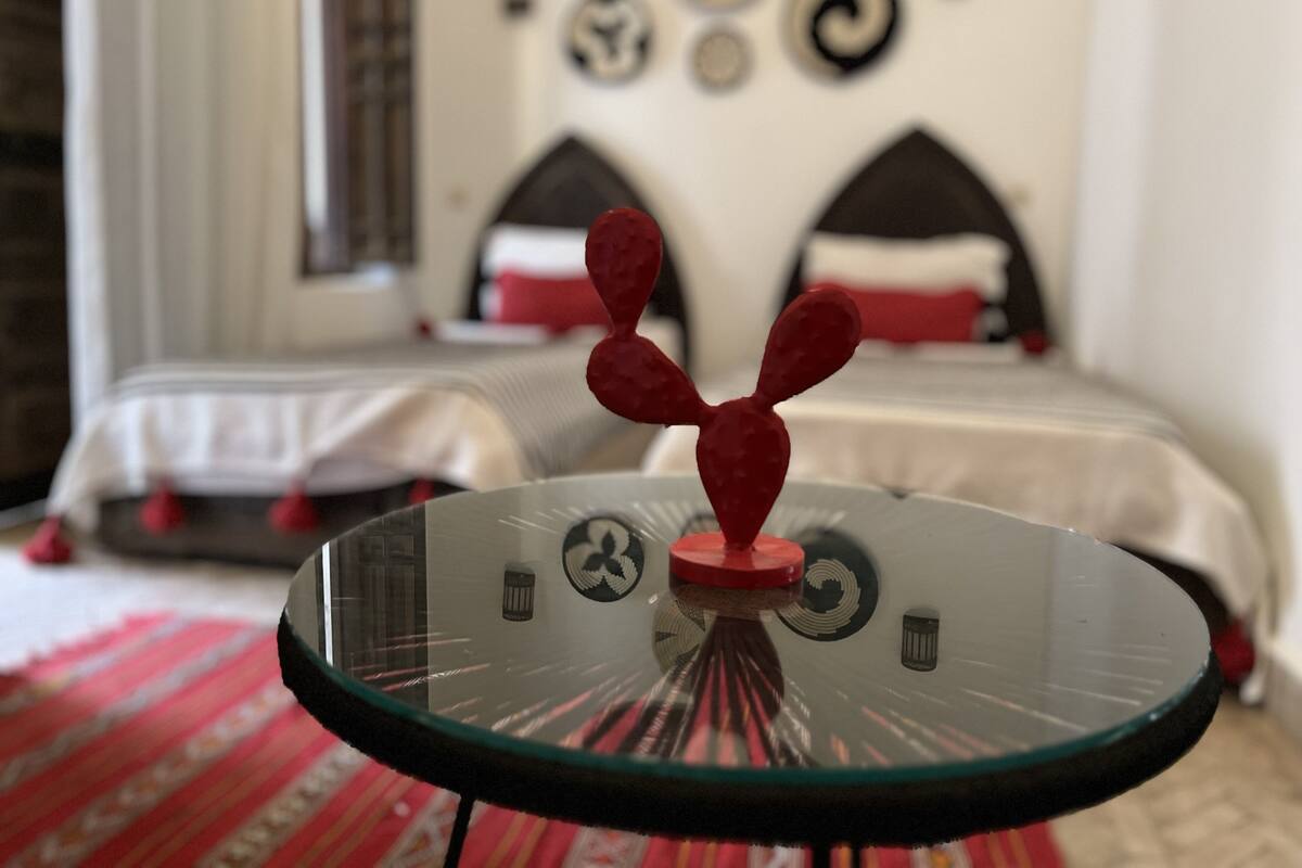 A room with a glass table in the foreground, a bed with a red and white bedspread in the background, and a wall with various decorative items.