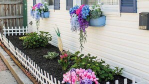 A white mobile home with a white picket fence, flower boxes on the windows, and a garden bed with various plants.