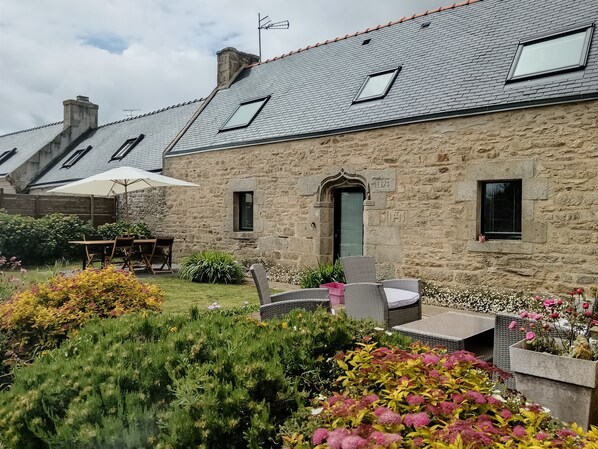 A stone building with a sloped roof, outdoor seating, and a garden with various plants and flowers.