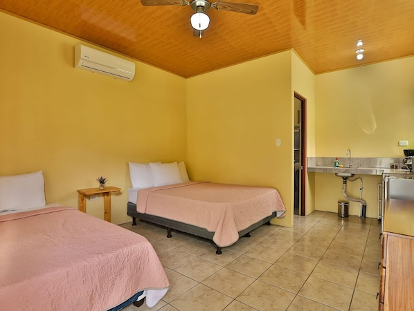 A room with two beds, a ceiling fan, an air conditioning unit, a small table with a plant, and a kitchenette area. - Chongos Place (La Fortuna)