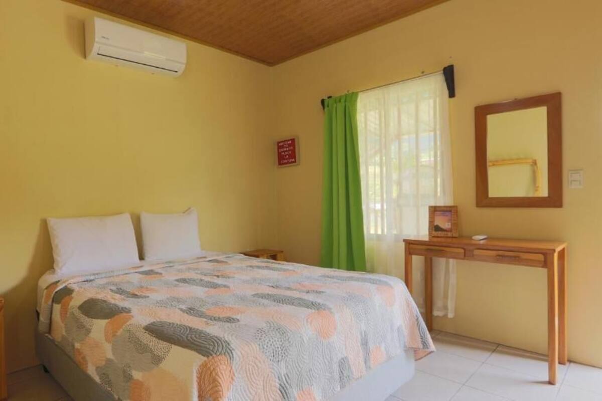 A bedroom with a bed, a bedside table, a desk with a mirror, and a window with curtains.