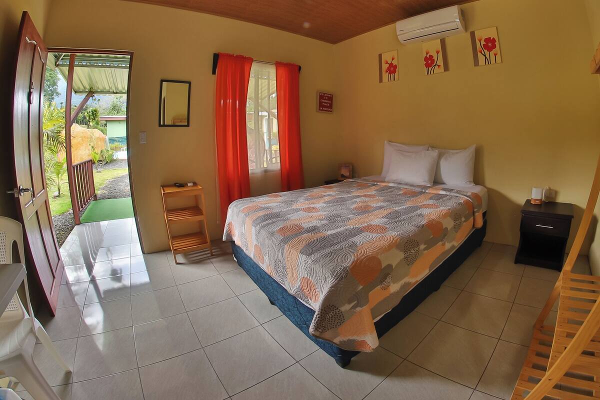 A bedroom with a bed, a nightstand, a wooden ladder, a small shelf, a mirror, and a door leading to a balcony.