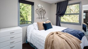 A bedroom with a bed, a dresser, two windows, and a view of greenery outside.