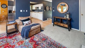 A living room with a sofa, a coffee table, a patterned rug, a wooden cabinet, and a kitchen in the background.