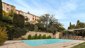 A swimming pool with a stone wall and greenery, a covered seating area, and a building in the background.