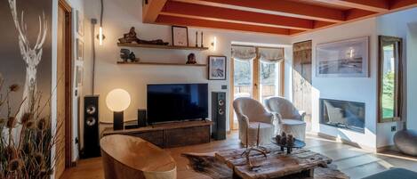 A modern living room with a wooden ceiling, a flat-screen TV, white chairs, a coffee table, and a fireplace.
