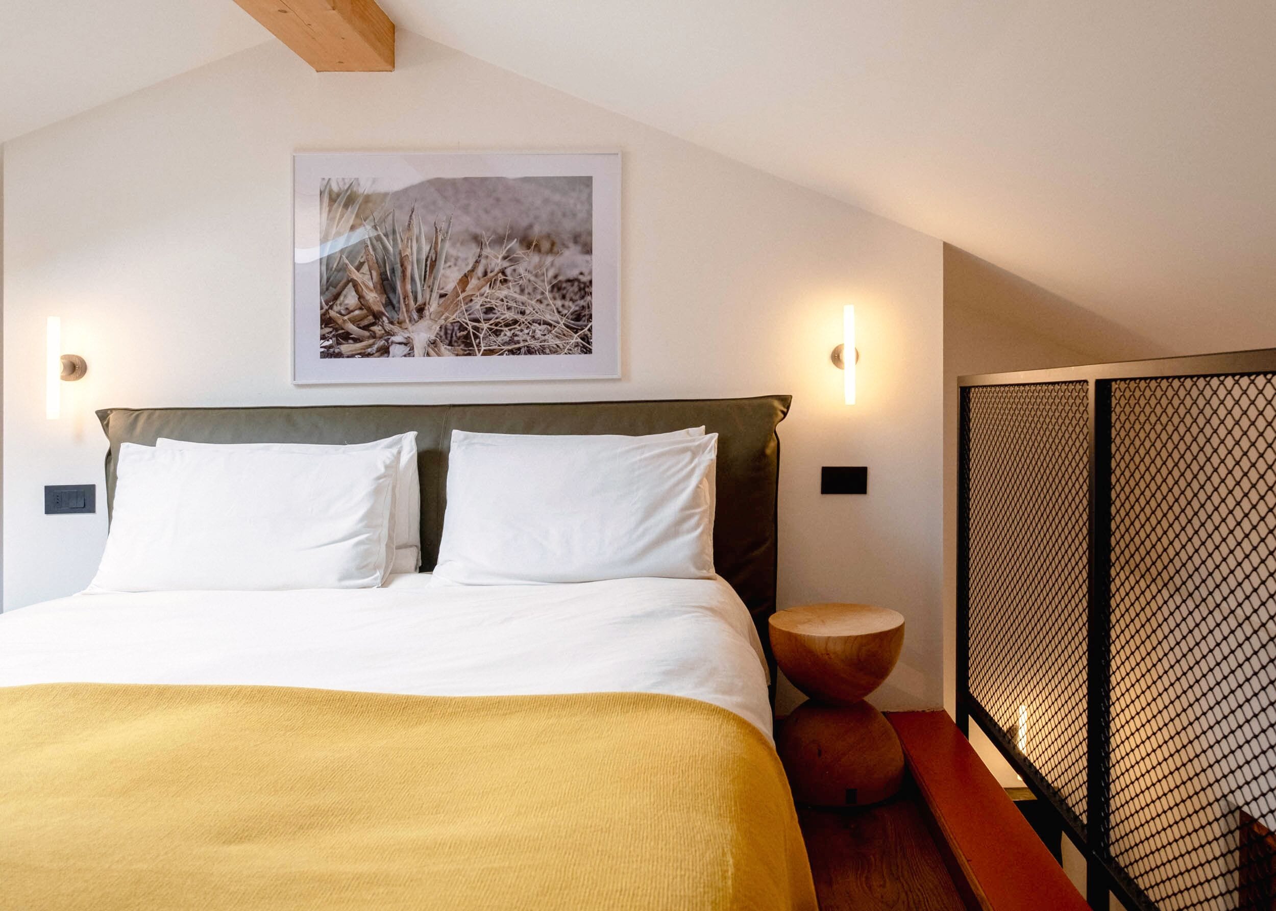 A bedroom with a bed, a headboard, a wall-mounted picture, two wall sconces, a round wooden stool, and a partition with a mesh design.