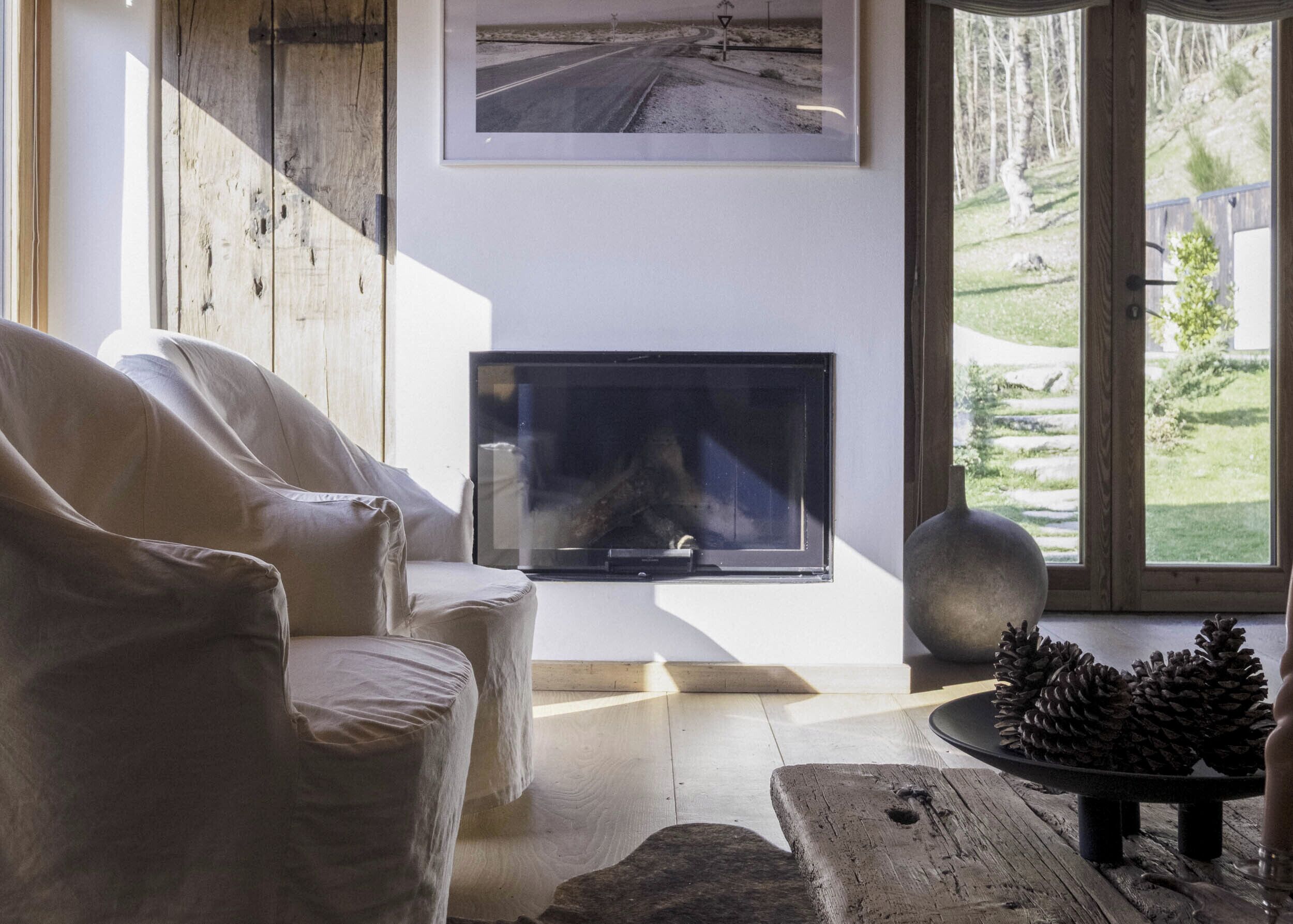 A living room with a fireplace, a large window with a view of a road and trees, and a rustic wooden table with pine cones.