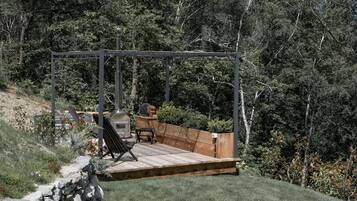 A wooden deck with a bench and a grill area, surrounded by a stone wall and lush greenery.