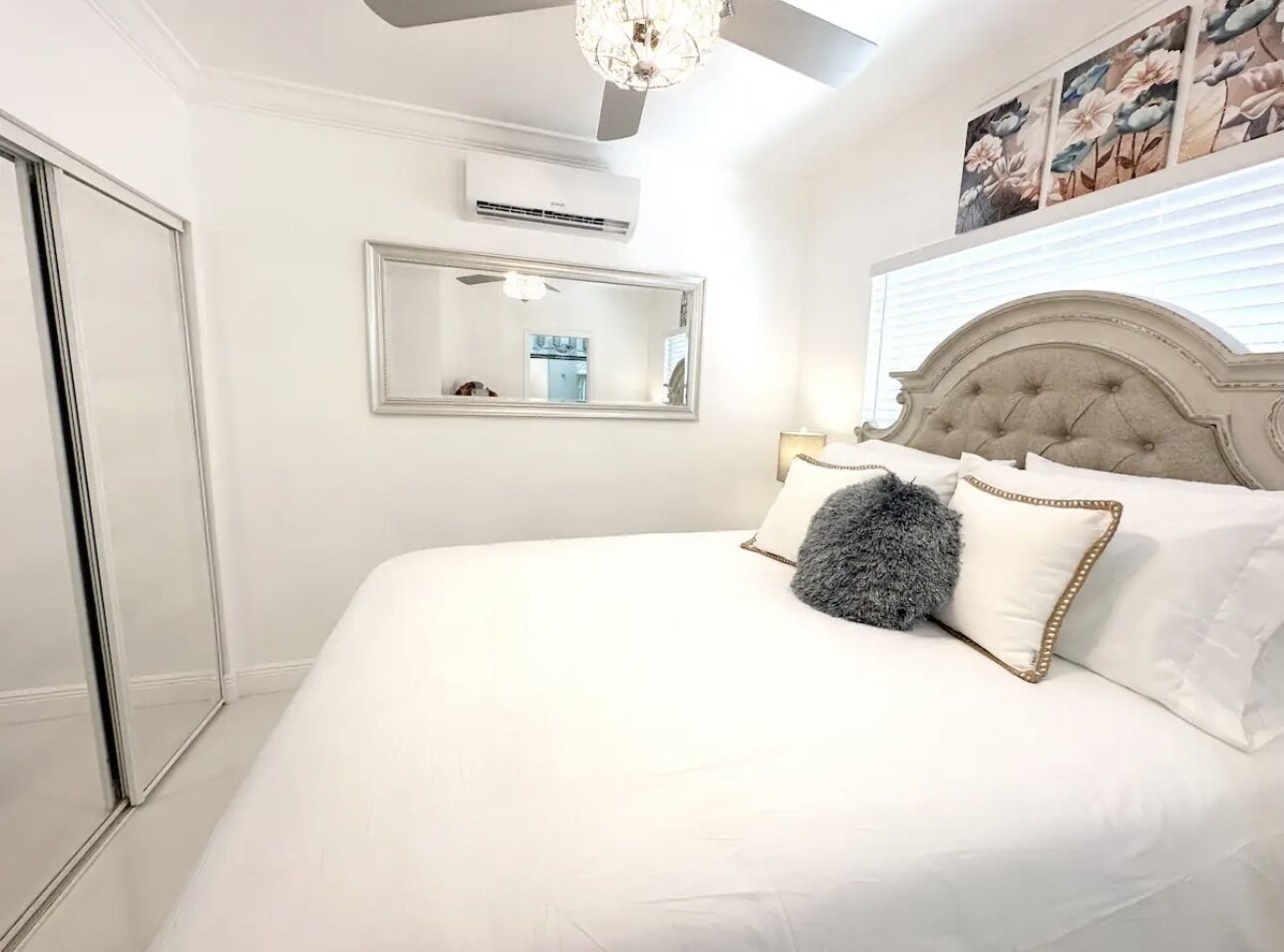 A bedroom with a large bed, a ceiling fan, a mirror, and a window with blinds.