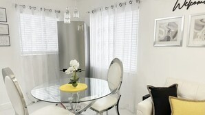 Un espace repas moderne avec une table en verre et des chaises blanches, un canapé blanc agrémenté de coussins jaunes et noirs, et un réfrigérateur en arrière-plan.