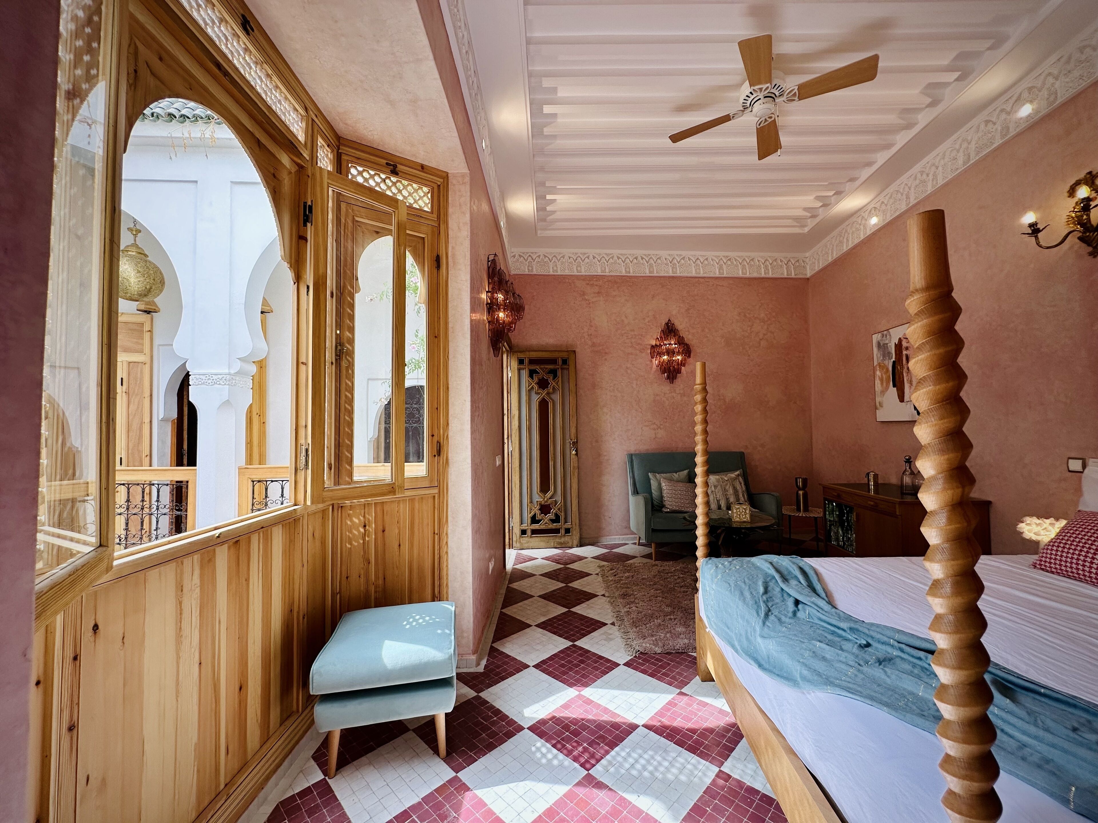 Una habitación con piso a cuadros, una cama con cabecera de madera, un banco, un ventilador de techo y vistas a un espacio exterior con arcos y un balcón.