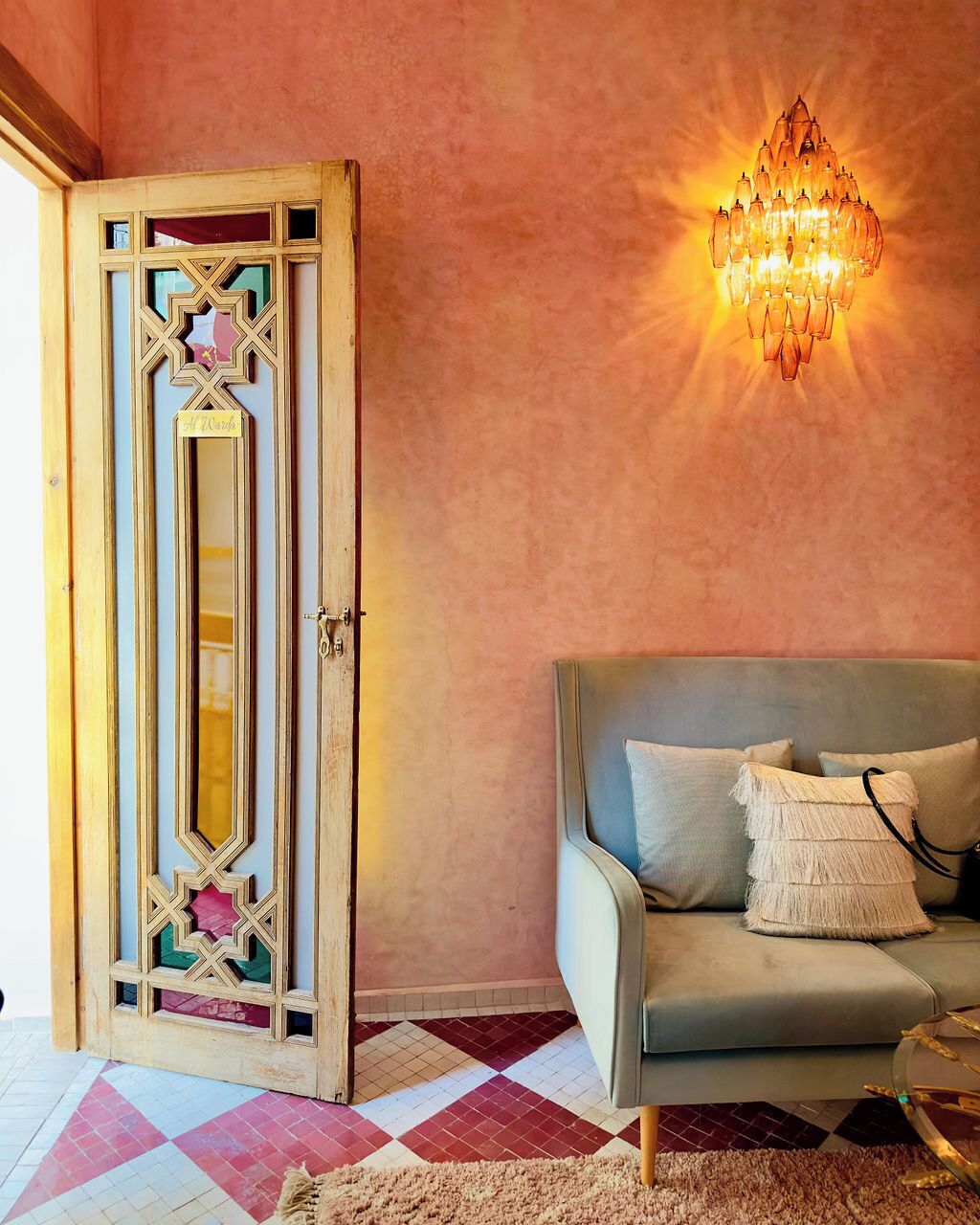 Una stanza con una porta in legno d'epoca, un divano grigio con cuscini, un tappeto decorato e un lampadario a parete.