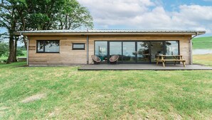 Ein modernes Haus mit Holzfassade, großen Glasfenstern und einer Terrasse mit zwei Korbstühlen und einem Holztisch.