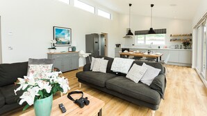 Ein modernes Wohnzimmer mit einem grauen Sofa, einem Holztisch und einer Vase mit weißen Blumen. Im Hintergrund ist eine Küche mit weißen Schränken und einem Essbereich zu sehen.