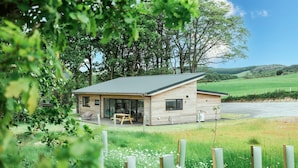 Eine Holzhütte mit geneigtem Dach, einer Terrasse mit Tisch und Stühlen und Blick auf ein grünes Feld und Bäume im Hintergrund.