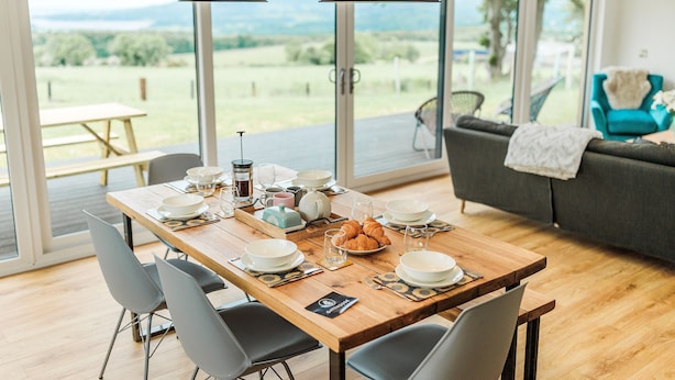 Ein Essbereich mit Holztisch, gedeckt für ein Frühstück, großen Fenstern mit Aussicht ins Freie und einer Couch im Hintergrund.