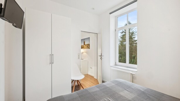 A bedroom with a bed, a TV, a window, and a door leading to a bathroom.