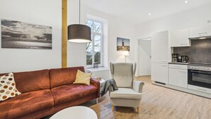 A modern living room with a brown leather sofa, a white coffee table, and a grey armchair. A large window provides natural light, and a kitchen with white cabinets and a stove is visible in the background.