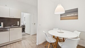 A modern kitchen with white cabinets, a built-in oven, and a sink. There's a dining area with a round white table and four white chairs. A pendant light hangs above the table, and a beach scene painting is on the wall.