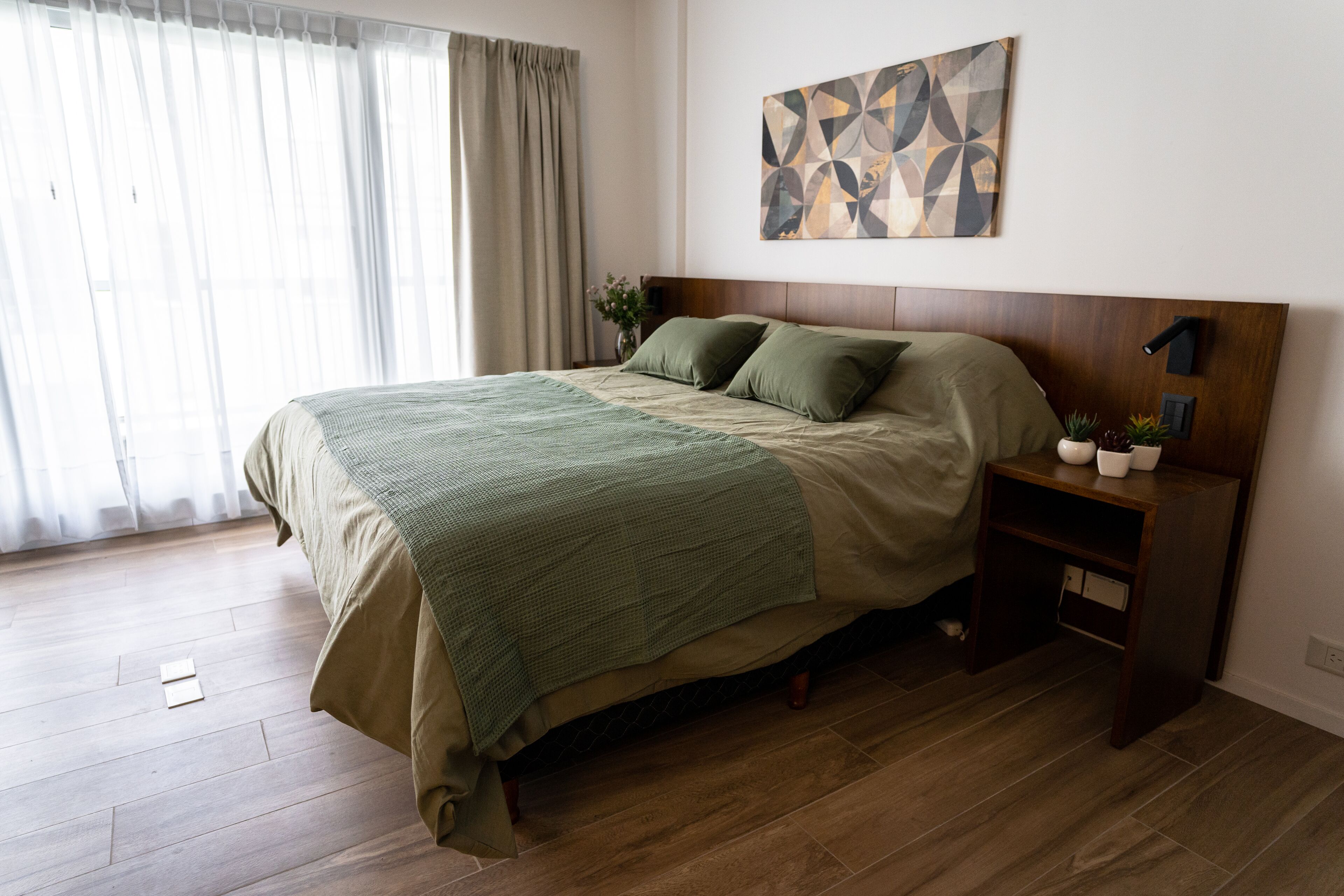 A bedroom with a wooden bed, a nightstand, a wall-mounted lamp, and a geometric artwork.