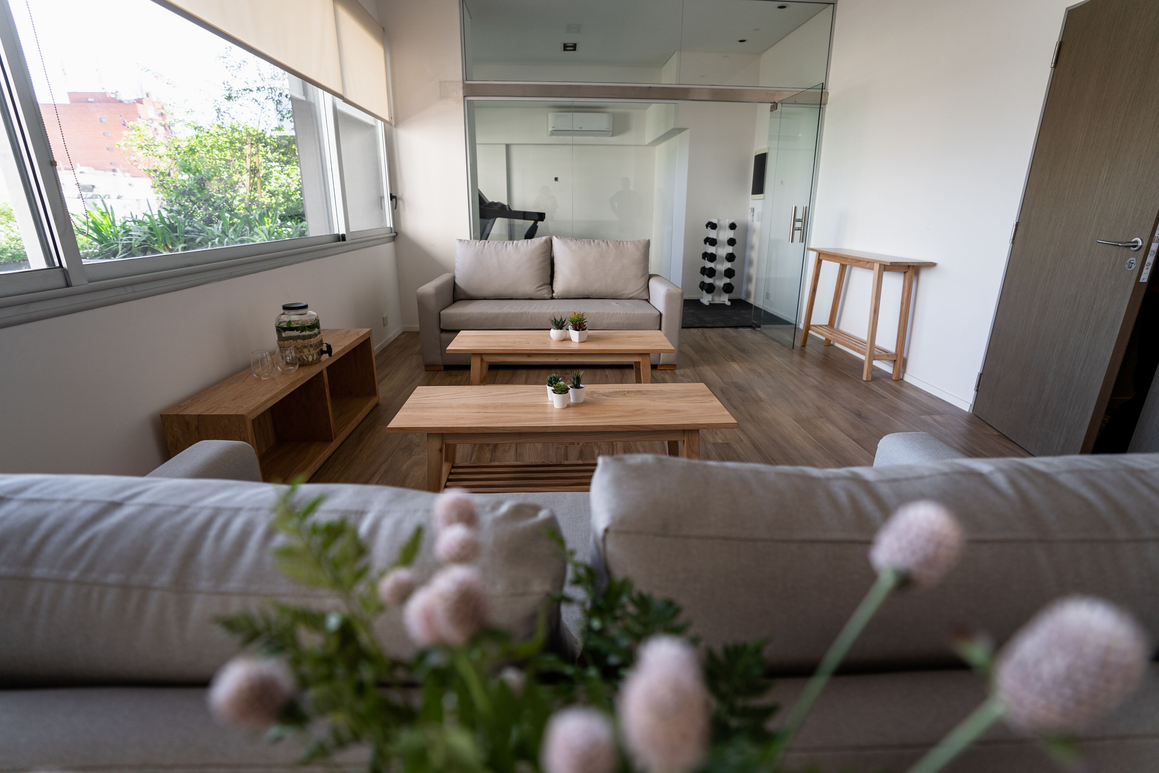 Un salon moderne avec un canapé, deux tables en bois et une vue sur un balcon, accessible par une porte vitrée.