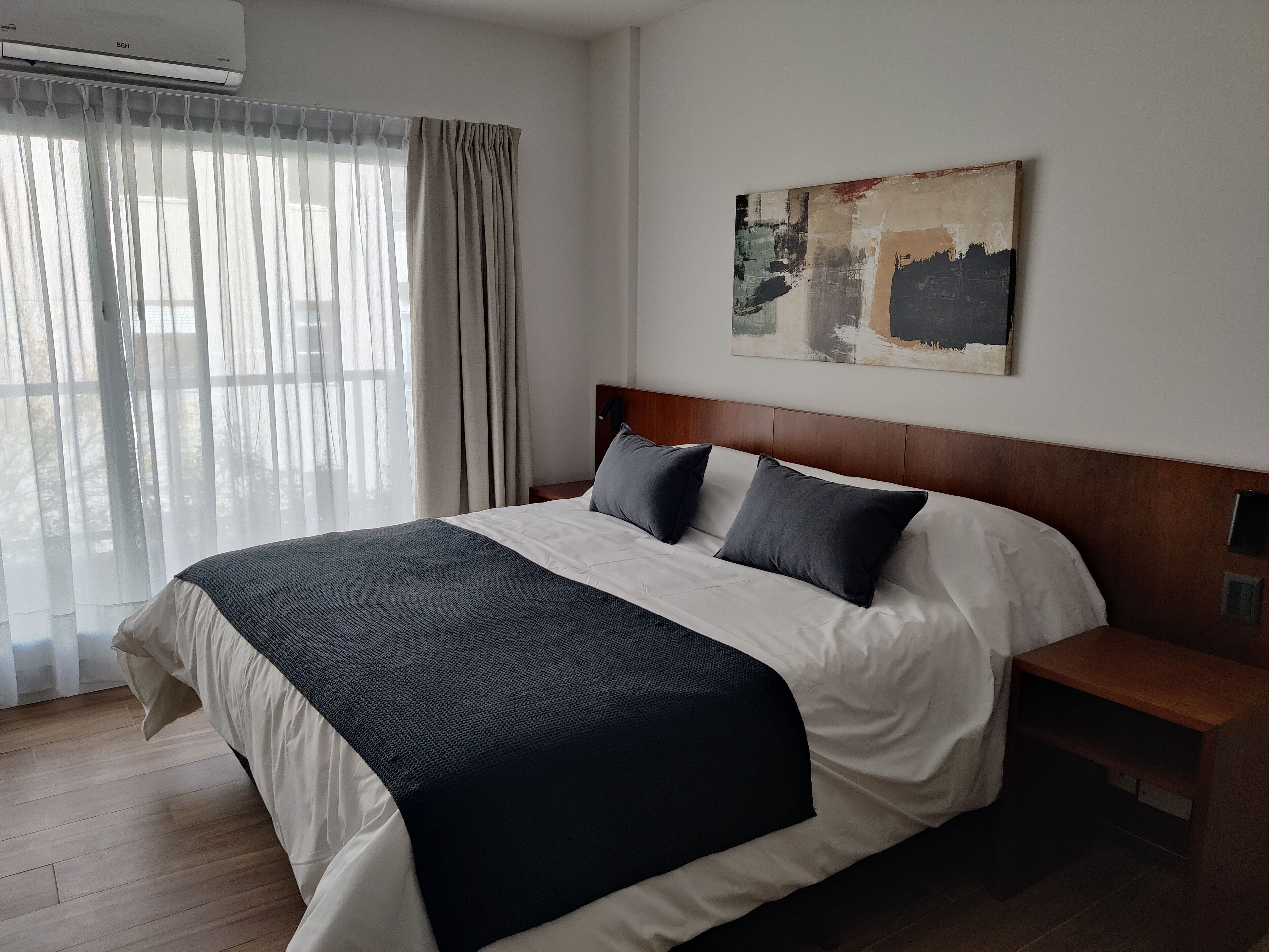 A neatly made bed with a dark headboard, a bedside table, and a large window with sheer curtains.