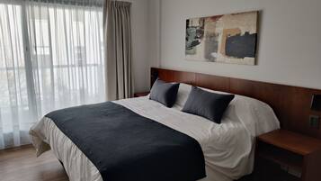 A neatly made bed with a dark headboard, a bedside table, and a large window with sheer curtains.