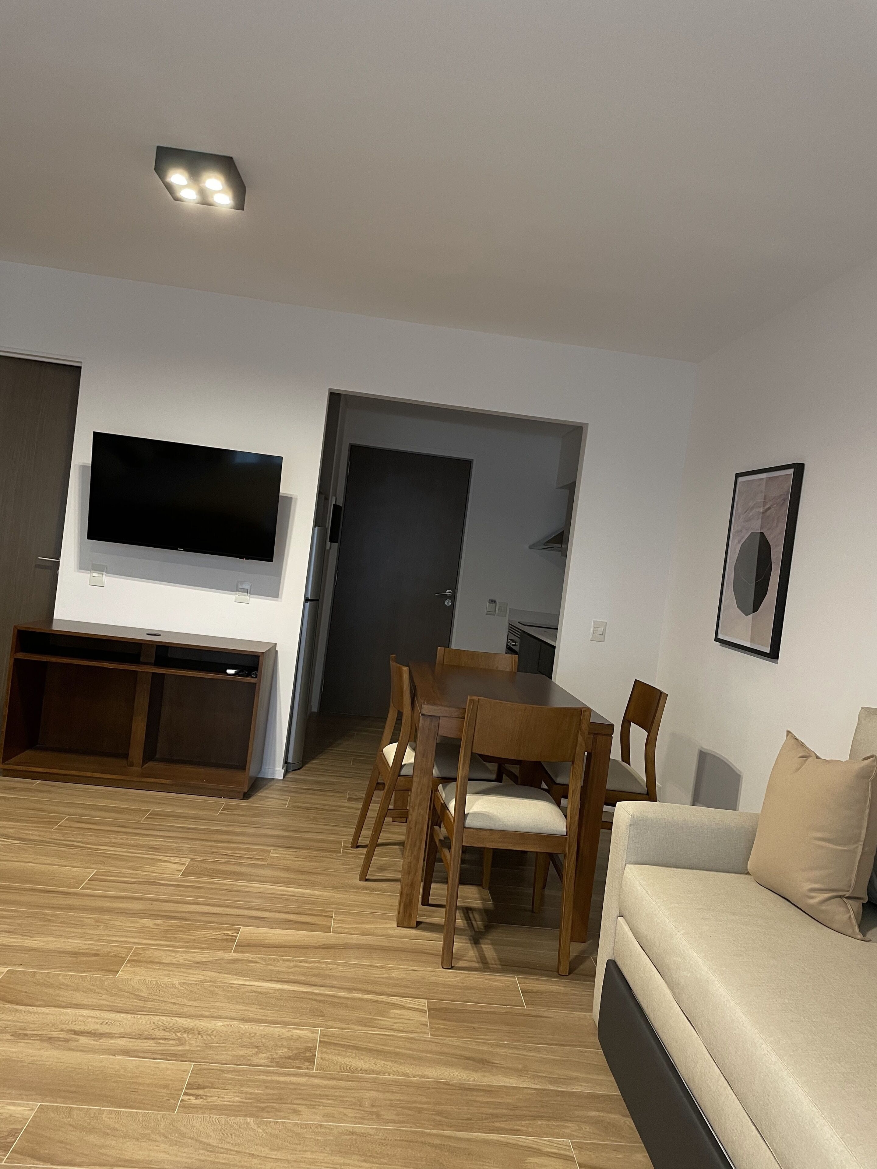 A modern living room with a flat-screen TV, wooden dining table, and chairs.