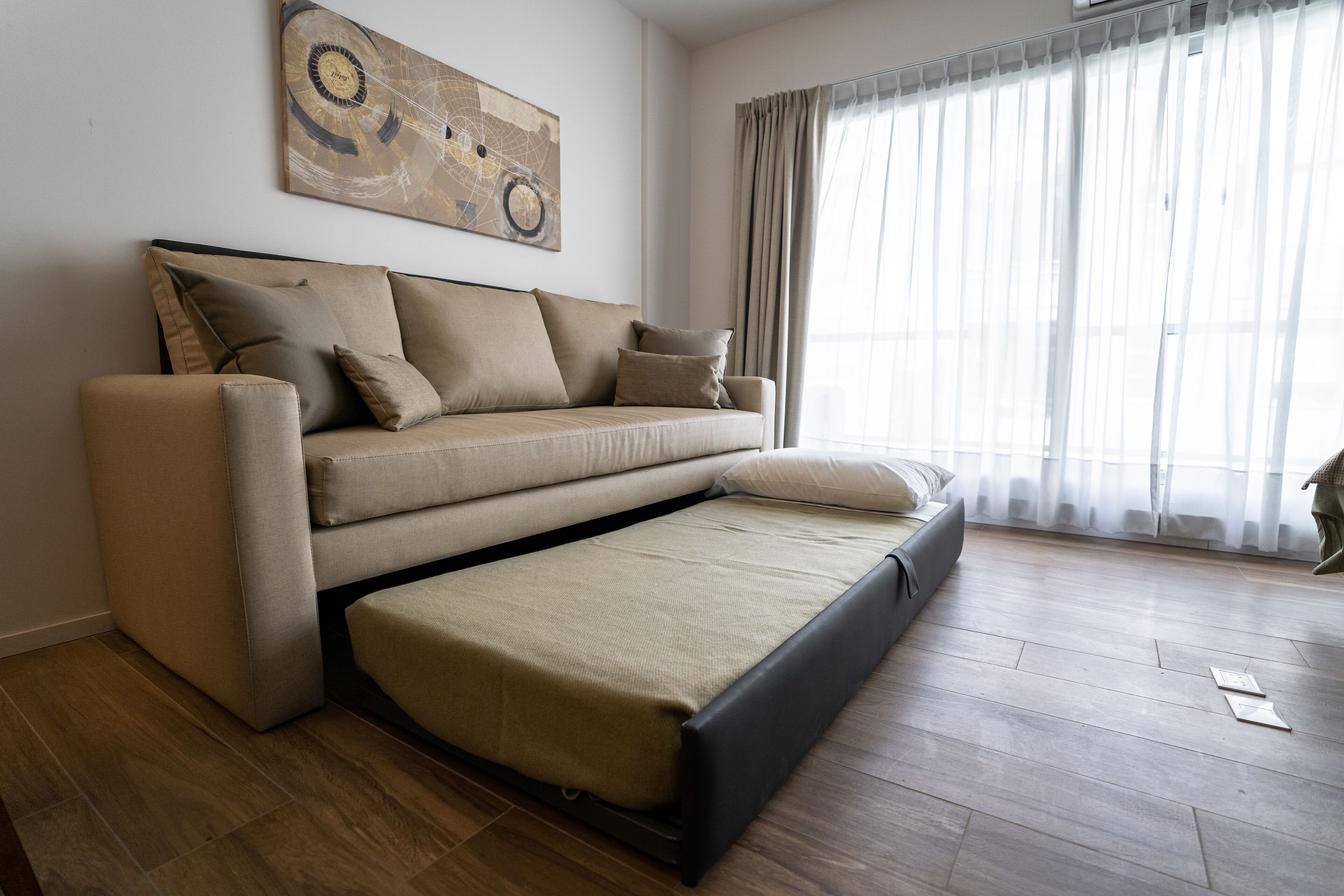 A modern living room with a sofa bed, a large abstract painting on the wall, and a large window with sheer curtains.