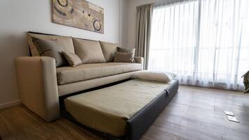 A modern living room with a sofa bed, a large abstract painting on the wall, and a large window with sheer curtains.