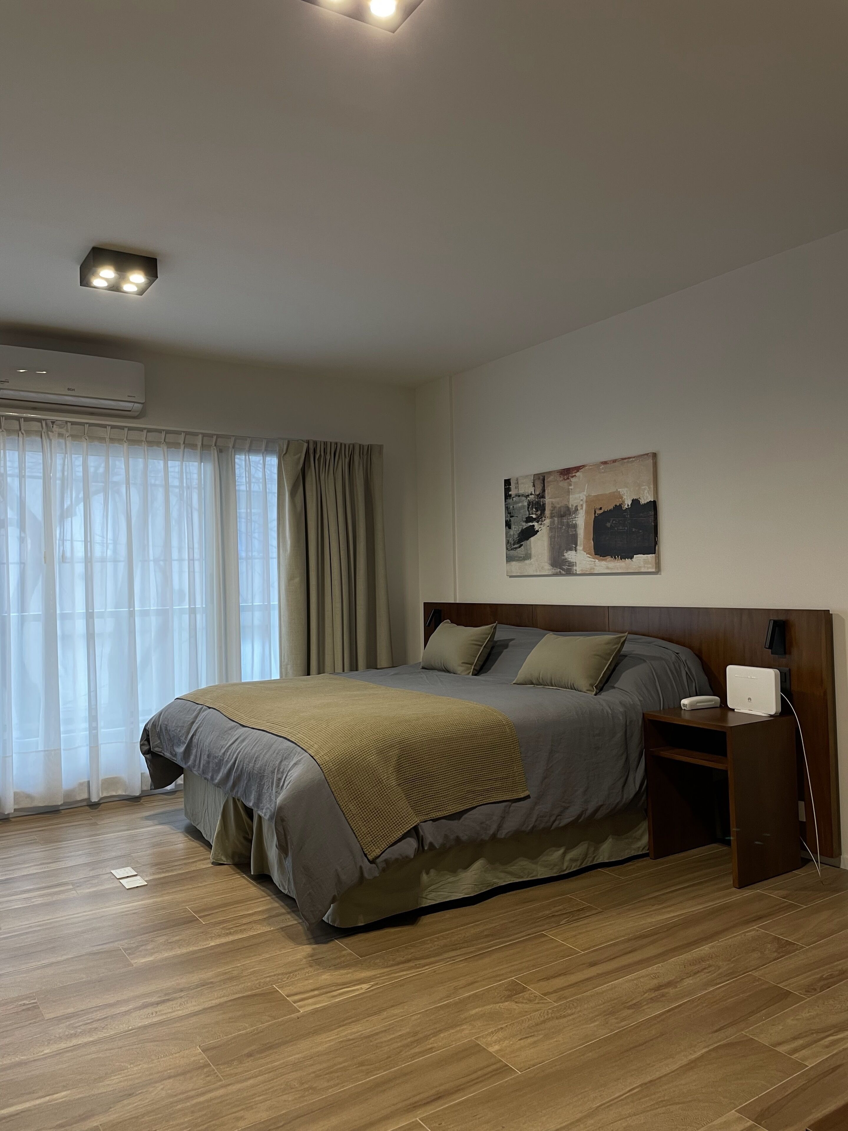 A bedroom with a bed, a bedside table with a lamp, an air conditioner, and a window with curtains.