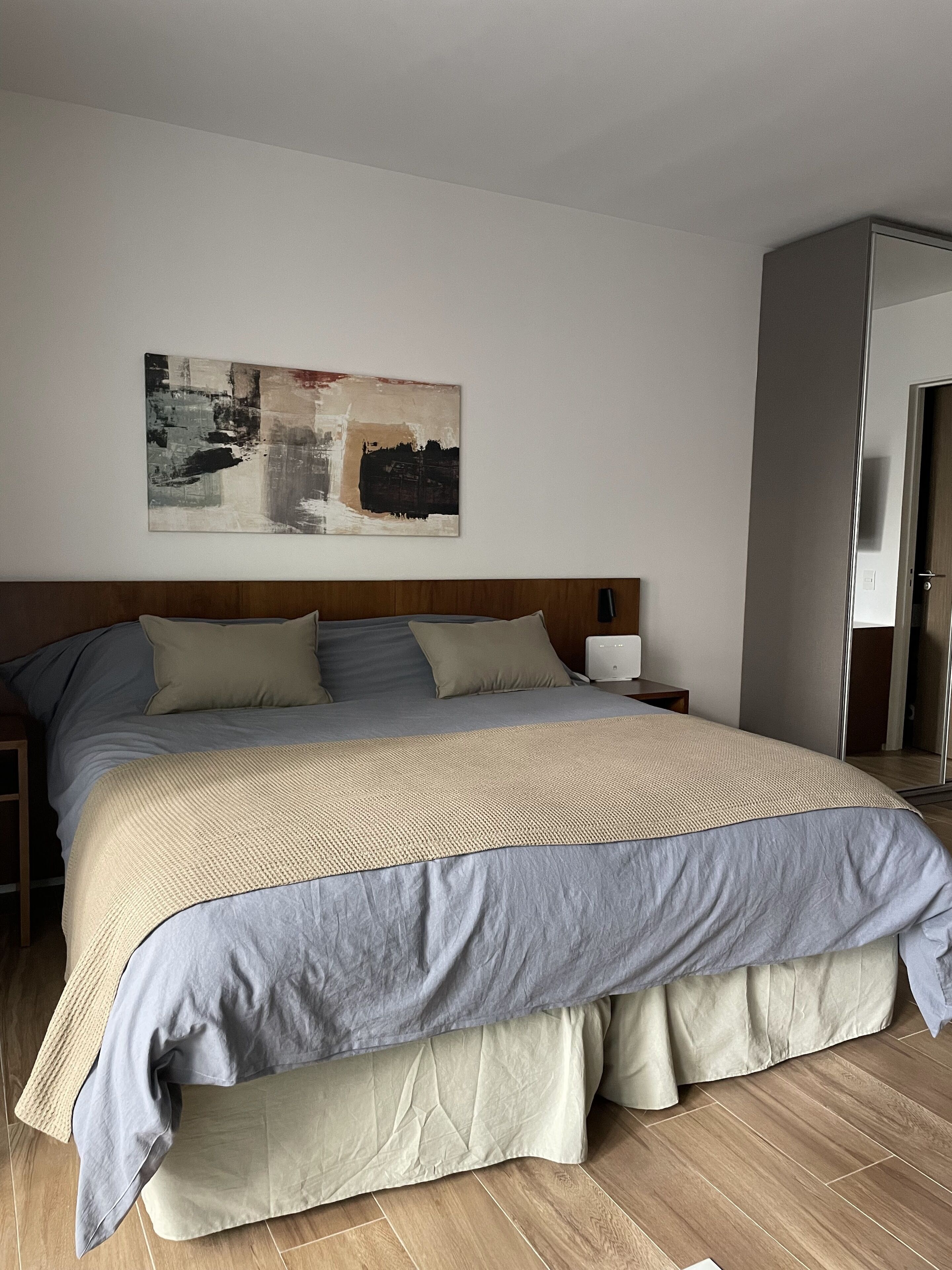 Un lit bien fait, avec une tête de lit en bois, une table de chevet sur laquelle se trouve une lampe, et un grand tableau abstrait accroché au mur.