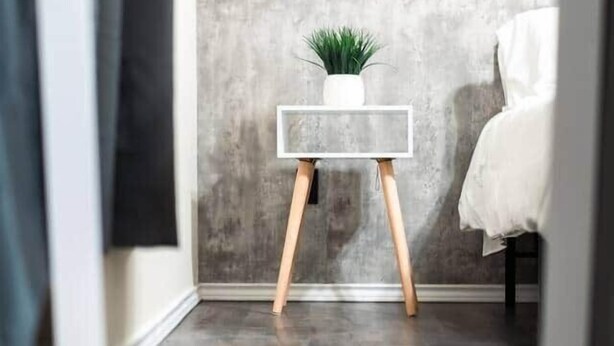 Une chambre moderne avec un mur en béton, une petite table de chevet blanche agrémentée d’une plante, et un lit blanc.