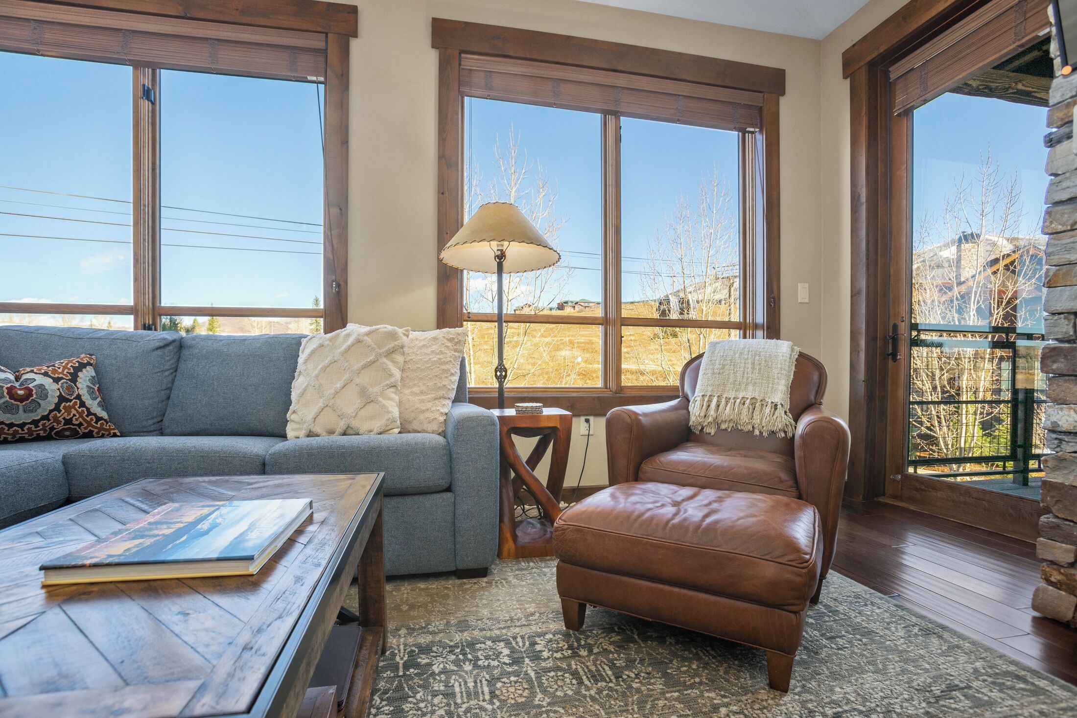 A cozy living room with a sofa, a leather chair, a coffee table with a book, and large windows offering a view of the outdoors.