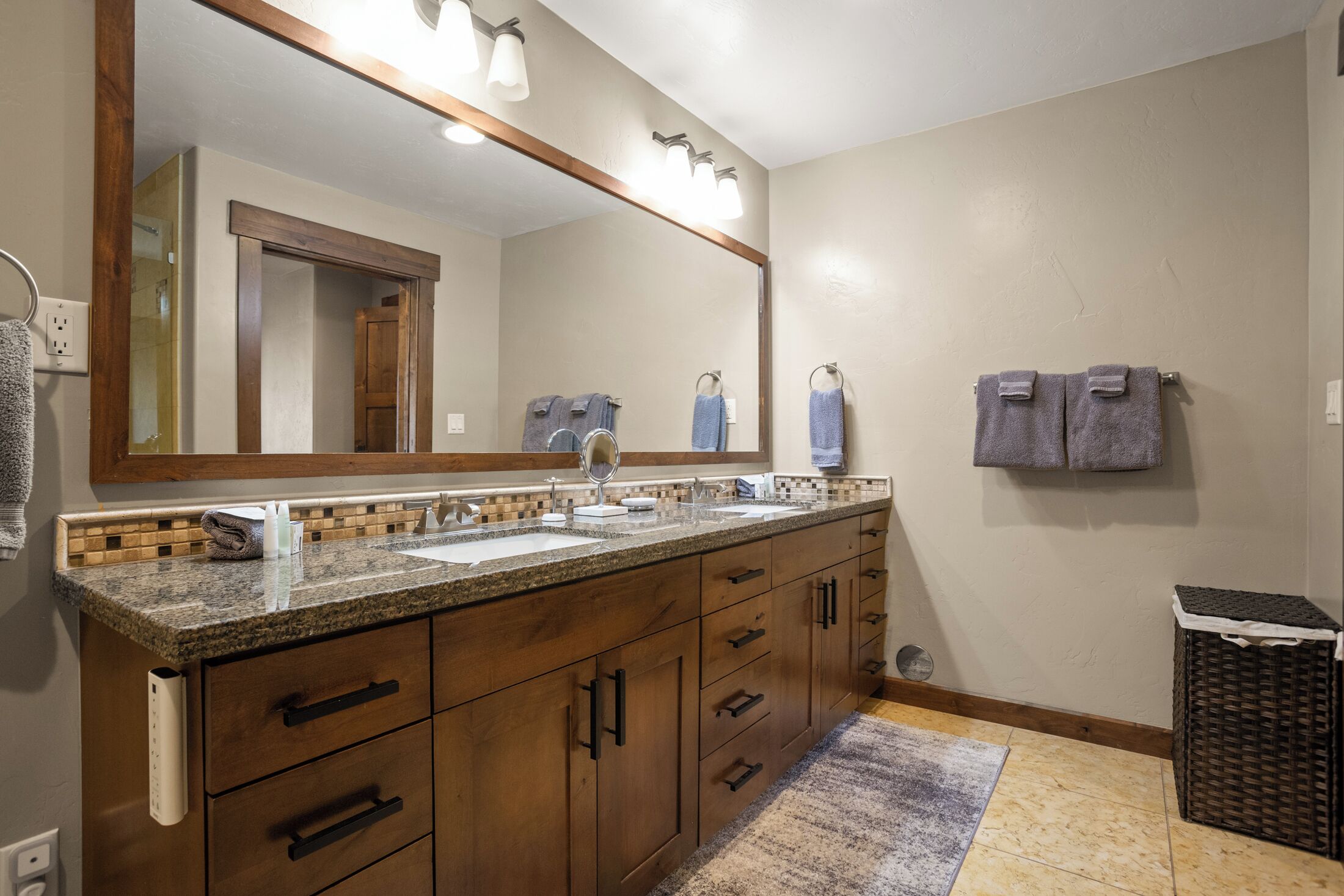 Un baño con tocador de madera, dos lavamanos, un espejo grande y un pequeño cubo de basura.